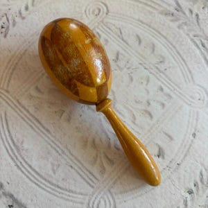 May include: A wooden maraca featuring a polished, oval-shaped head with a glossy finish and a geometric diamond pattern in alternating shades of light and dark brown. The instrument includes a smooth, slender, light-colored wooden handle.