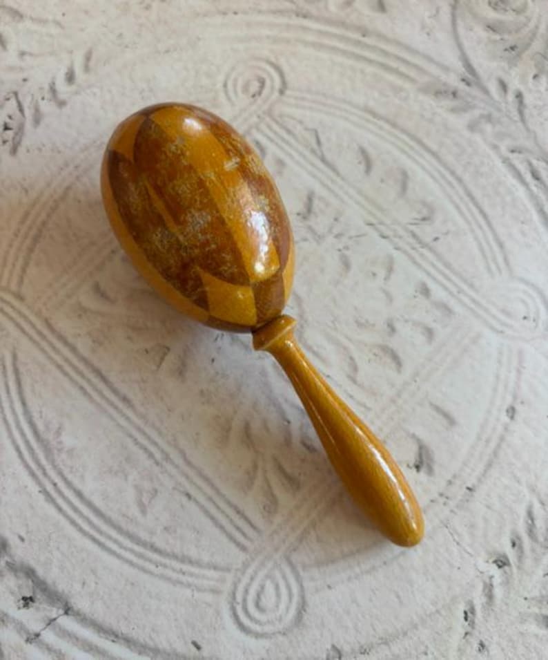 May include: A wooden maraca featuring a polished, oval-shaped head with a glossy finish and a geometric diamond pattern in alternating shades of light and dark brown. The instrument includes a smooth, slender, light-colored wooden handle.