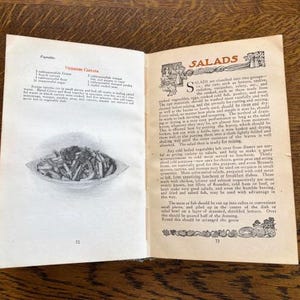 May include: Open vintage cookbook with a recipe for "Viennese Carrots" on the left page and a section on "Salads" on the right page. The left page includes a black and white illustration of a bowl of carrots. The right page has a decorative border with a floral design.