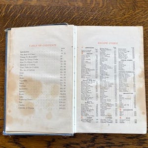 May include: An open vintage cookbook with a table of contents and recipe index. The book has a cream-colored paper cover with red and black text.