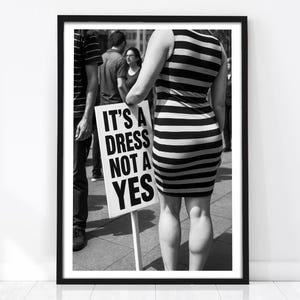 Könnte beinhalten: Ein schwarz-weiß gerahmter Druck, der eine Person in einem ärmellosen Kleid mit horizontalen Streifen zeigt, die ein rechteckiges Schild mit dem Text IT'S A DRESS NOT A YES hält. Die Szene zeigt einen öffentlichen Ort mit weiteren Personen.