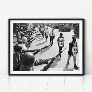 May include: A black and white photograph showing a line of individuals wearing helmets and holding rifles pointed towards a group of people walking in a protest. The protesters carry signs that clearly display the text I AM A MAN in bold letters.