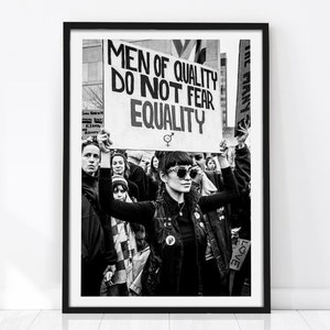 Peut inclure: Une impression encadrée en noir et blanc représentant une scène de manifestation. Une personne au premier plan porte des vêtements sombres et des lunettes de soleil, tenant une pancarte indiquant : MEN OF QUALITY DO NOT FEAR EQUALITY.