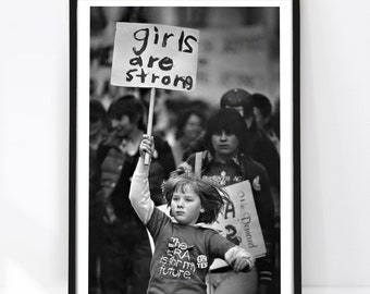 May include: A black and white framed photograph depicting a protest scene. A child in the foreground holds a sign that reads "girls are strong" and wears a shirt with the text "the ERA is for my future." Other protesters are visible behind.
