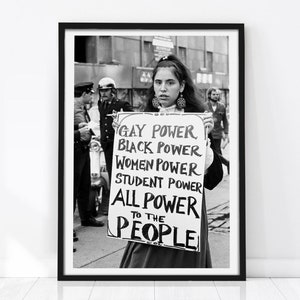 May include: A black and white framed photograph showing a person holding a sign that reads: GAY POWER, BLACK POWER, WOMEN POWER, STUDENT POWER, ALL POWER TO THE PEOPLE. The scene includes figures in the background and a motorcycle in the street.