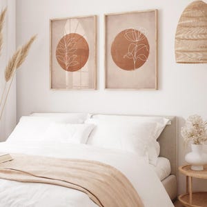 Op de afbeelding: Een slaapkamer met een bed met wit beddengoed en een perzikkleurige deken. Twee ingelijste prints met abstracte botanische ontwerpen hangen boven het bed. Een geweven lampenkap en vaas dragen bij aan de inrichting.