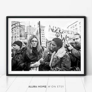 Black and white photograph of Gloria Steinem at a women’s rights protest, surrounded by activists, signs, and photographers in a city square.