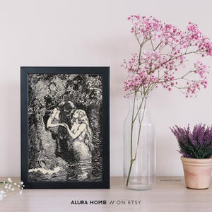 May include: A framed black and white print depicting a mermaid and a person in a forest setting. The print is displayed on a light-colored surface, next to a vase of pink flowers and a small potted plant. The text "ALURA HOME // ON ETSY" is at the bottom.