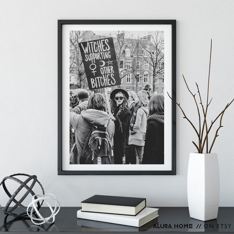 K&ouml;nnte beinhalten: Schwarz-Wei&szlig;-Druck in einem Rahmen, der eine Menschenmenge zeigt. Eine Person in der Mitte h&auml;lt ein Schild mit der Aufschrift "Witches Supporting Other Bitches", einem Venus-Symbol und einem Stern. Der Druck wird auf einer schwarzen Oberfl&auml;che pr&auml;sentiert.