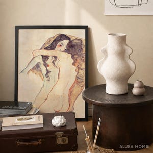 K&ouml;nnte beinhalten: Ein gerahmter Kunstdruck mit zwei sich umarmenden Figuren, eine gro&szlig;e, strukturierte, cremewei&szlig;e Keramikvase mit geschwungener Silhouette, eine kleine runde beige Vase, ein brauner Vintage-Koffer, ein B&uuml;cherstapel und ein kleines wei&szlig;es Deko-Objekt.