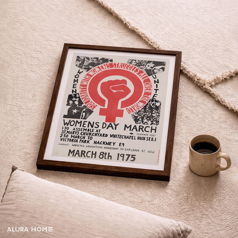 Puede incluir: Un p&oacute;ster enmarcado con marco de madera oscura. El p&oacute;ster presenta un s&iacute;mbolo de pu&ntilde;o rojo y el texto "WOMENS DAY MARCH MARCH 8th 1975". Una taza beige con caf&eacute; est&aacute; a la derecha. El p&oacute;ster est&aacute; sobre una superficie beige.
