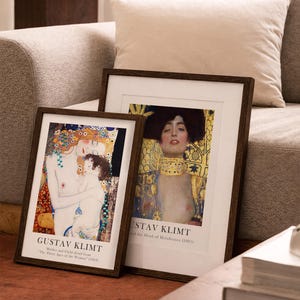 May include: Two framed art prints featuring Gustav Klimt paintings. The left print shows Mother and Child from The Three Ages of Woman, and the right print shows Judith and the Head of Holofernes. Both have dark wood frames and white matting.