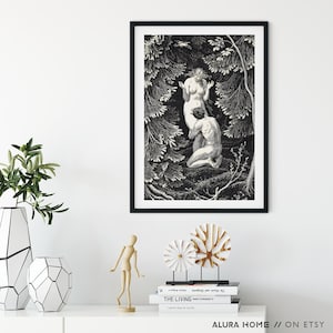 May include: Black framed art print depicting a classical scene of two nude figures in a forest setting. The artwork is in black and white, with detailed shading and a vintage aesthetic. The print is displayed on a white surface with decorative objects.