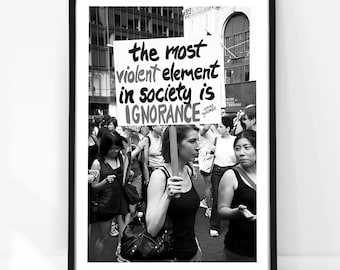 May include: A black and white framed print featuring a protest scene with a person holding a sign that reads, "the most violent element in society is IGNORANCE -emma goldman." The image captures a crowd in an urban setting with architectural details.