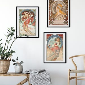 Puede incluir: Tres l&aacute;minas art&iacute;sticas enmarcadas con ilustraciones al estilo de Alphonse Mucha de mujeres con prendas fluidas y fondos circulares complejos, expuestas en marcos negros sobre una pared blanca encima de un banco de madera con una manta gris.