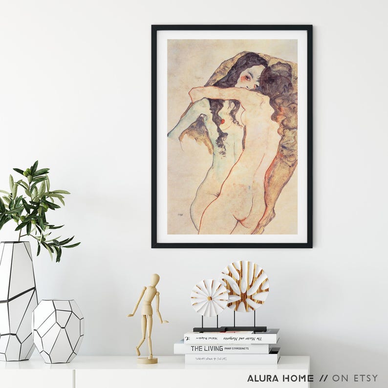 May include: Framed art print featuring a nude figure study in muted tones. The artwork, set in a black frame, depicts two figures embracing. The print is displayed on a white surface with decorative objects and greenery.