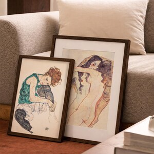 May include: Two framed art prints with dark wooden frames. One print features a seated figure in green and black, the other depicts two figures embracing. The prints are displayed on a red-brown surface.