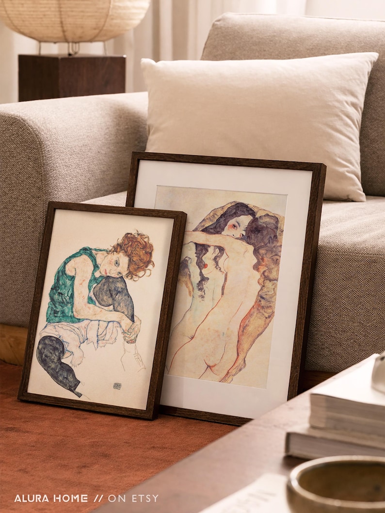 May include: Two framed art prints with dark wooden frames. One print features a seated figure in green and black, the other depicts two figures embracing. The prints are displayed on a red-brown surface.