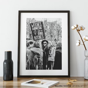 K&ouml;nnte beinhalten: Schwarz-Wei&szlig;-Druck in einem Rahmen, der eine Menschenmenge zeigt. Eine Person h&auml;lt ein Schild mit der Aufschrift "Witches Supporting Other Bitches". Das Bild ist eine Fotografie einer Demonstration.