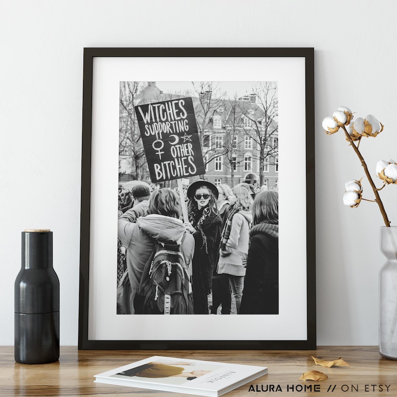 K&ouml;nnte beinhalten: Schwarz-Wei&szlig;-Druck in einem Rahmen, der eine Menschenmenge zeigt. Eine Person h&auml;lt ein Schild mit der Aufschrift "Witches Supporting Other Bitches". Das Bild ist eine Fotografie einer Demonstration.