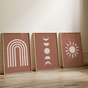 May include: Three rectangular wall art prints in light wooden frames, each featuring a terracotta background with white graphic illustrations. The left print shows a rainbow, the center shows moon phases, and the right shows a sunburst design.