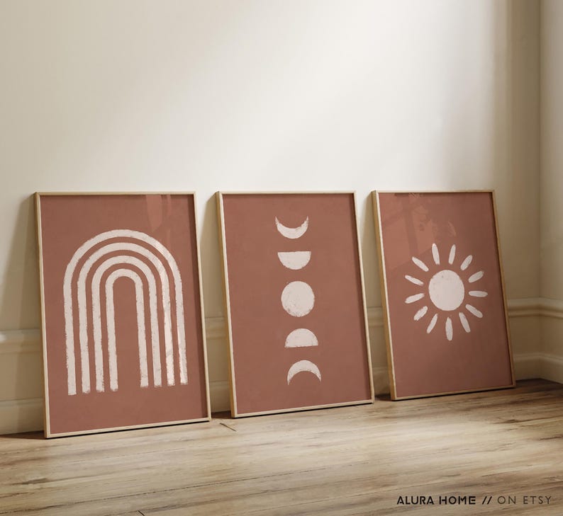 May include: Three rectangular wall art prints in light wooden frames, each featuring a terracotta background with white graphic illustrations. The left print shows a rainbow, the center shows moon phases, and the right shows a sunburst design.