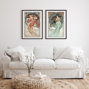 Puede incluir: Dos impresiones art&iacute;sticas enmarcadas con ilustraciones de estilo Art Nouveau de mujeres con cabello y vestidos fluidos. La obra de arte tiene una est&eacute;tica vintage con una paleta de colores de rojos, verdes y azules. Las impresiones se exhiben sobre un sof&aacute; blanco.