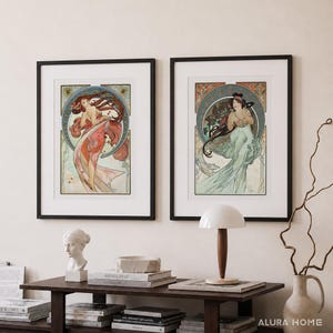 Puede incluir: Dos impresiones art&iacute;sticas enmarcadas que presentan ilustraciones de estilo Art Nouveau de mujeres con cabello y vestimenta fluidos. Las impresiones, enmarcadas en negro, presentan un dise&ntilde;o circular con elementos florales y geom&eacute;tricos. La obra de arte se exhibe sobre una mesa de madera oscura.