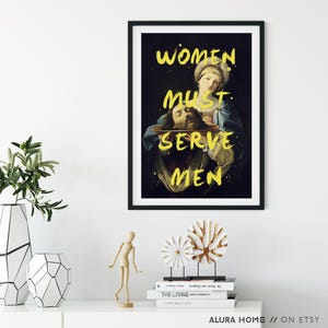May include: A framed art print with a dark background and yellow text that reads "WOMEN MUST SERVE MEN." The artwork depicts a classical painting of a woman holding a platter with a severed head. The print is displayed on a white shelf.
