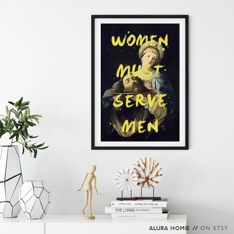 May include: A framed art print with a dark background and yellow text that reads "WOMEN MUST SERVE MEN." The artwork depicts a classical painting of a woman holding a platter with a severed head. The print is displayed on a white shelf.