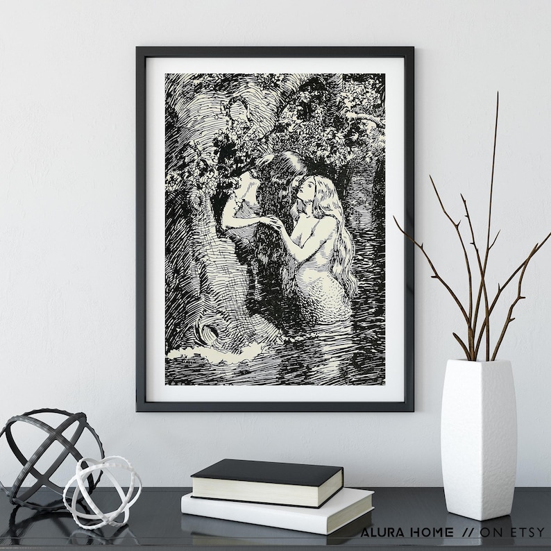 May include: Black and white framed art print featuring a detailed illustration of two mermaids in a watery environment. One mermaid is looking up, while the other is reaching out. The art print is displayed on a black surface with decorative items.