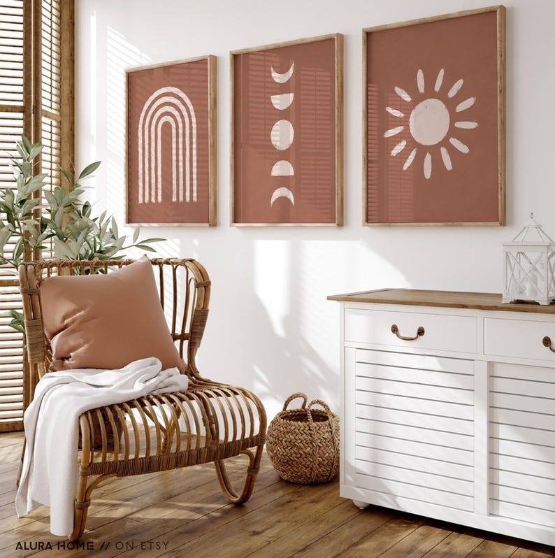 May include: Three framed wall art prints featuring white line drawings on a terracotta background. The left print shows a rainbow, the center print displays moon phases, and the right print depicts a sun. The frames are light-colored wood.