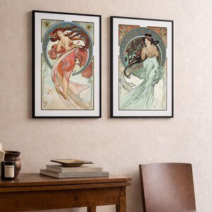Puede incluir: Dos impresiones art&iacute;sticas enmarcadas con ilustraciones de estilo Art Nouveau de mujeres con cabello y prendas fluidas. La obra de arte tiene una paleta de colores suaves con detalles en rojo, naranja y verde, sobre un fondo crema.