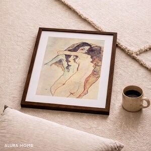 May include: A framed print of a nude figurative artwork with a dark brown wooden frame. The artwork depicts two figures embracing, rendered in muted tones. A beige mug with coffee sits nearby, alongside a pillow.