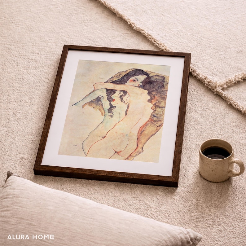 May include: A framed print of a nude figurative artwork with a dark brown wooden frame. The artwork depicts two figures embracing, rendered in muted tones. A beige mug with coffee sits nearby, alongside a pillow.