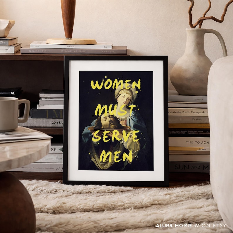 May include: A framed art print with the text "WOMEN MUST SERVE MEN" in yellow over a classical painting. The artwork features a woman holding a platter with a man's head. The print is in a black frame.