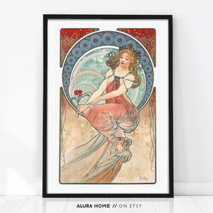 Puede incluir: Una l&aacute;mina art&iacute;stica enmarcada que muestra la ilustraci&oacute;n de una persona con cabello suelto y una prenda rosa drapeada, sosteniendo una flor roja. El dise&ntilde;o incluye un borde circular azul y blanco con detalles florales rojos y la firma Mucha.
