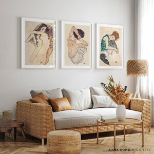 May include: Three framed art prints featuring nude figures in various poses. The prints are hung on a white wall above a wicker sofa with brown throw pillows.
