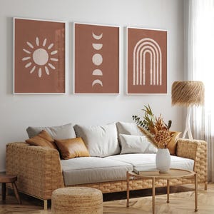 May include: Three framed wall art prints featuring white line drawings on a terracotta background. The left print shows a sun, the center print displays moon phases, and the right print depicts a rainbow arch. The text ALURA HOME // ON ETSY is visible.
