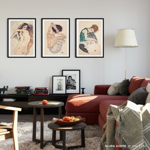 May include: Three framed black and white art prints of Egon Schiele's drawings hanging on a white wall above a red sectional sofa. The prints feature a woman with long hair, a woman sitting with her arms crossed, and a woman sitting with her legs crossed.