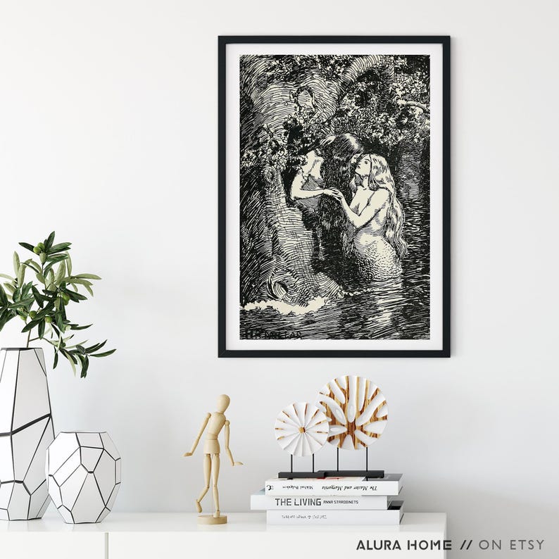 May include: Black framed art print featuring a detailed black and white illustration of two mermaids in a watery environment, surrounded by foliage. The artwork is displayed on a white wall, with decorative items in the foreground.