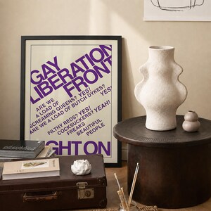 May include: A framed poster with purple text reading GAY LIBERATION FRONT, ARE WE A LOAD OF SCREAMING QUEENS? YES! ARE WE A LOAD OF BUTCH DYKES? YES! FILTHY REDS? YES! COCKSUCKERS? YEAH! FREAKS BEAUTIFUL PEOPLE RIGHT ON, next to a textured off-white vase.