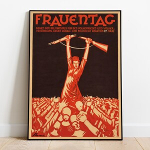 May include: A framed vintage poster with a black border. The poster features a red and beige illustration with German text. A central figure holds up weapons, surrounded by raised hands. The text at the top reads "FRAUENTAG".