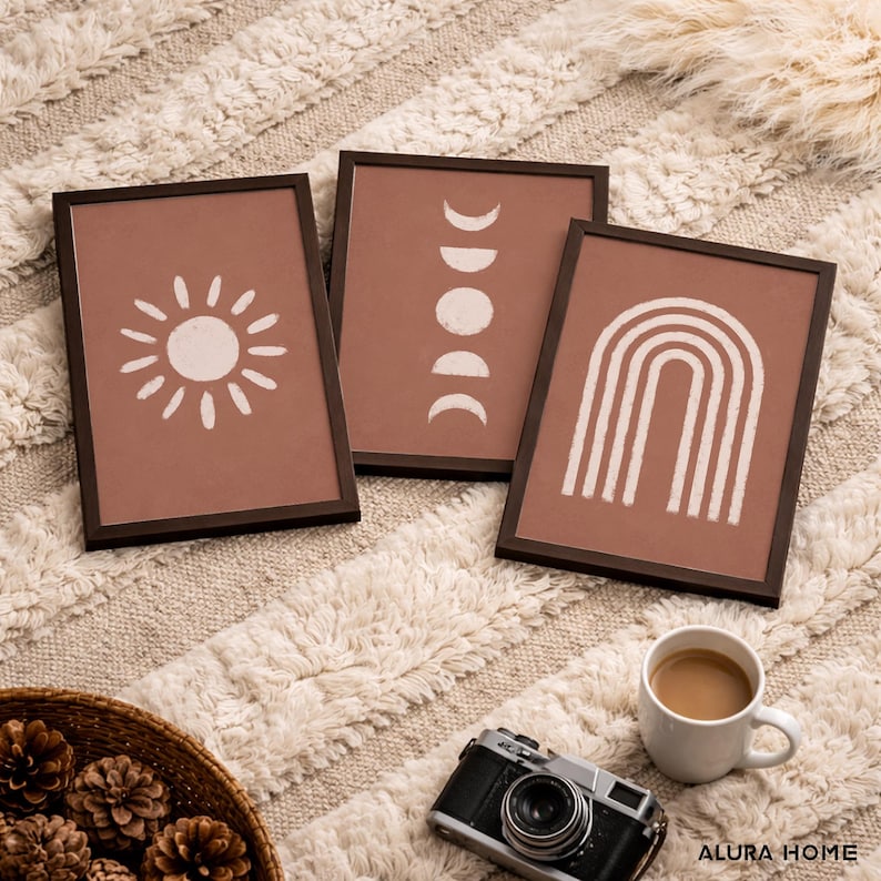May include: Three rectangular wall art prints in dark brown frames. The prints feature white line art on a terracotta background, depicting a sun, moon phases, and a rainbow. The text Alura Home is visible in the bottom right corner of the image.