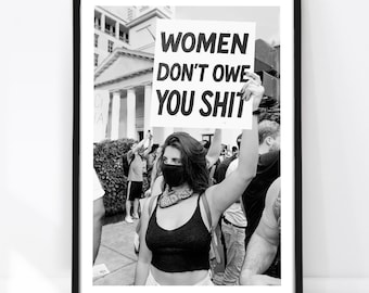 May include: A black and white framed print featuring a person wearing a black face mask and a dark tank top, holding a white protest sign with the bold black text WOMEN DON'T OWE YOU SHIT, set against a backdrop of a public outdoor gathering.