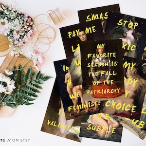 May include: A collection of art prints with yellow text on dark backgrounds. The prints feature classical paintings with overlaid phrases like "My favorite season is the fall of the patriarchy." Floral arrangements and decorative items surround the prints.