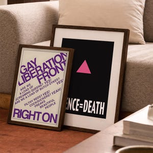 May include: Two framed posters with dark wood frames. The left poster has a white background with purple text reading GAY LIBERATION FRONT, RIGHT ON, and other phrases. The right poster has a black background with a pink triangle and SILENCE=DEATH.