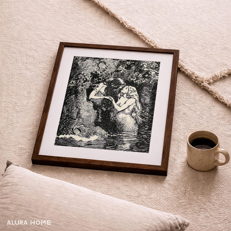 May include: A framed black and white illustration of mermaids in a forest setting. The artwork is in a dark brown wooden frame. A beige mug with coffee sits to the right of the frame. The image is on a beige rug.