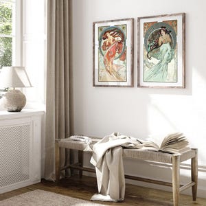 Puede incluir: Dos impresiones art&iacute;sticas enmarcadas con ilustraciones de estilo Art Nouveau de mujeres con cabello suelto y fondos florales ornamentados. Las impresiones est&aacute;n en marcos de madera y cuelgan de una pared blanca. Un banco con una parte superior tejida y una manta est&aacute;n frente a la pared.