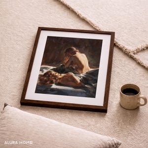 Pode incluir: Uma obra de arte emoldurada com uma moldura de madeira marrom escura. A obra retrata duas figuras em uma cama, renderizadas em tons quentes. Uma caneca de cer&acirc;mica bege com l&iacute;quido escuro est&aacute; &agrave; direita da moldura. Uma almofada bege clara est&aacute; em primeiro plano.